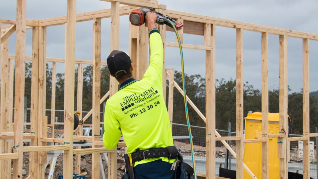 A construction worker in a bright yellow shirt operates a power tool on a timber frame at a building site. The background displays construction materials and a bushland area under an overcast sky, with support by Smart Apprenticeship Solutions.