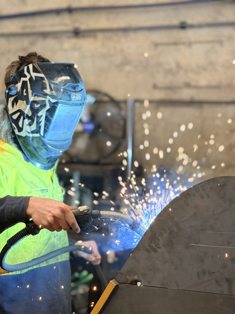 In a workshop, a person in a patterned welding helmet and neon safety jacket welds metal, with support by Smart Apprenticeship Solutions.