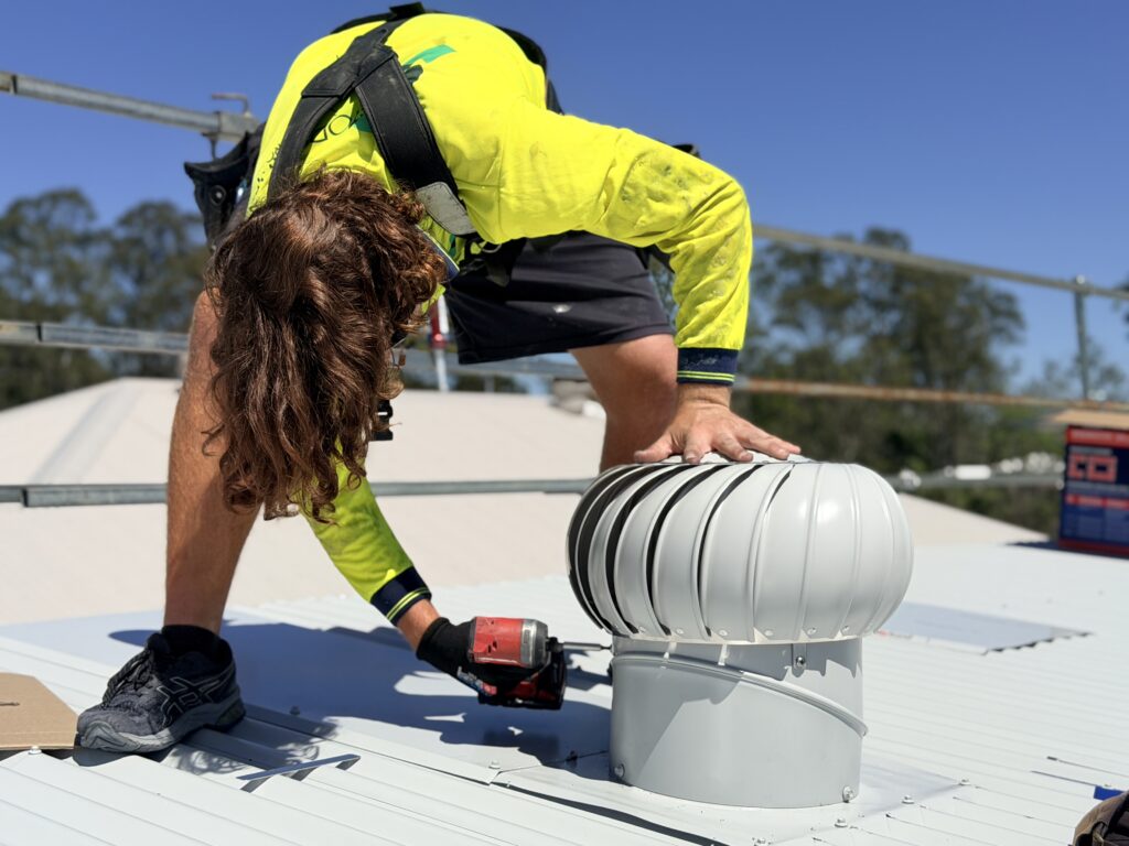 A worker in a high-vis shirt and safety harness fits a silver roof vent to a corrugated metal roof with a power drill, supported by Smart Apprenticeship Solutions. Blue sky and trees appear in the background.