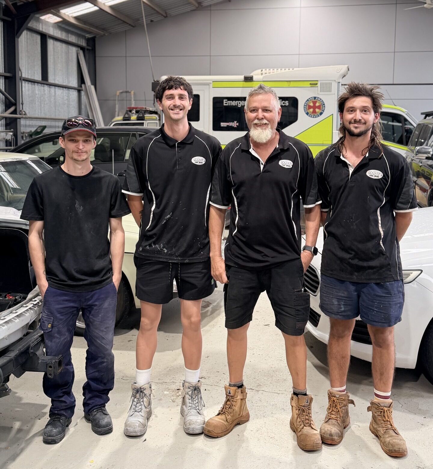 Four men smile inside a workshop with cars and an ambulance, supported by Smart Apprenticeship Solutions. Their uniforms vary slightly.