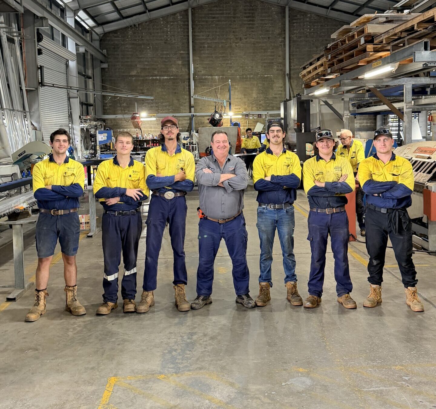 Seven workers in yellow and navy uniforms stand with arms crossed in a warehouse, supported by Smart Apprenticeship Solutions.
